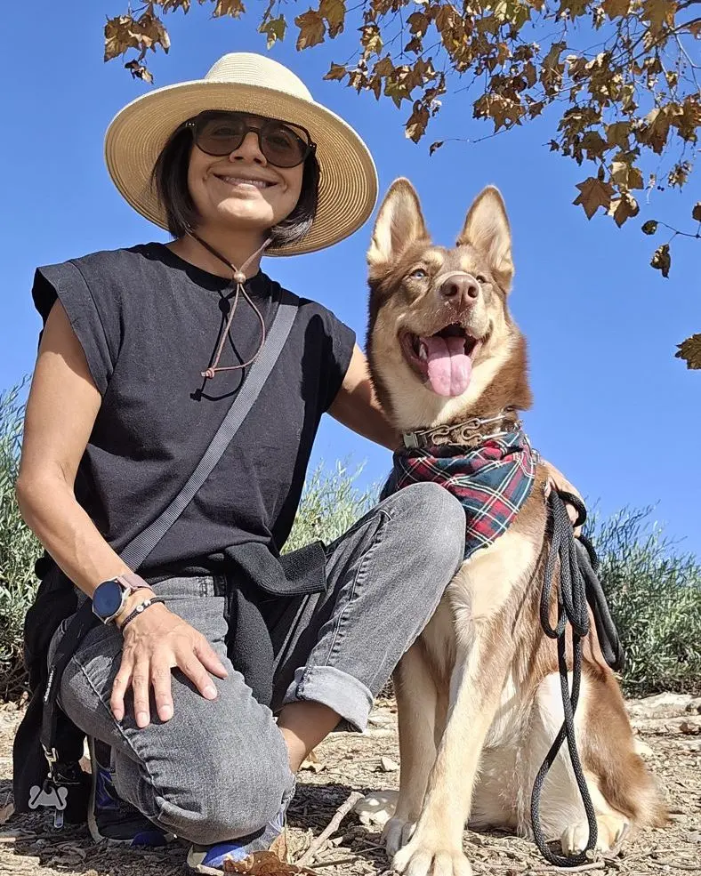 Angelica kneeling beside a smiling shepherd mix wearing a plaid bandana on a sunny trail